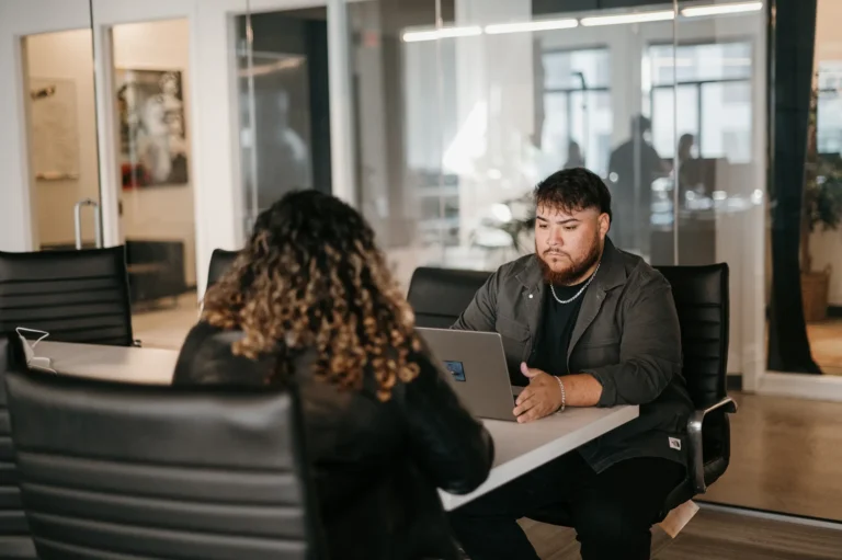 In a well-lit office with large glass partitions, two individuals sit across from each other at a conference table. One person is focused on the laptop, expertly navigating website design. This environment fosters both creativity and productivity.
